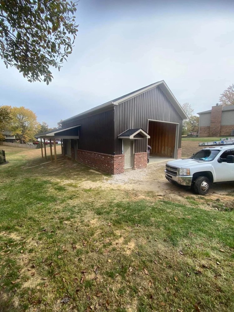 Completed pole barn with dark metal siding, brick skirt, and entry door awning